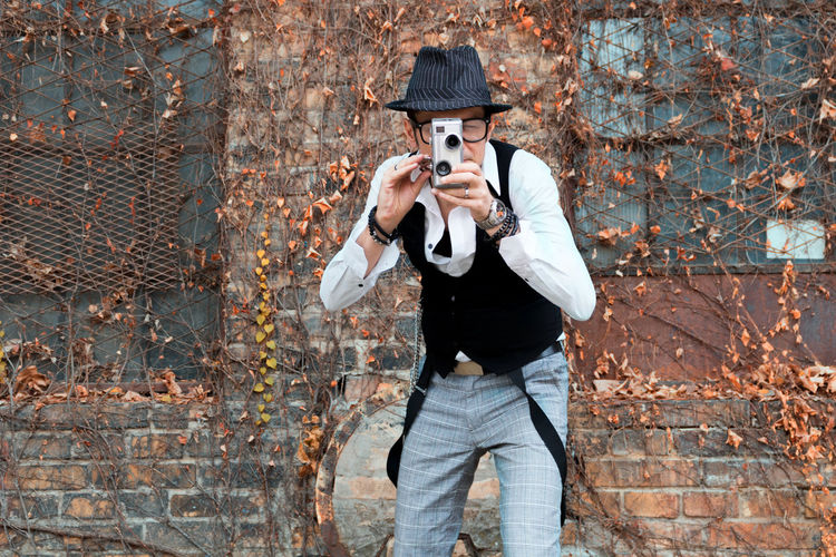 Mature man photographing while standing against brick wall during autumn