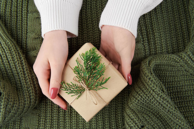 Girls hands holding a gift with fir decoration on knitted green sweater. cozy christmas still life