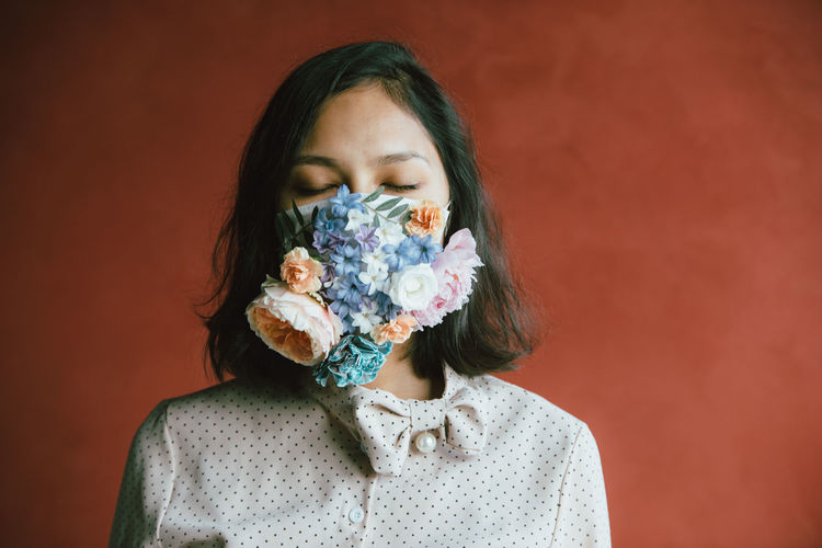 PORTRAIT OF YOUNG WOMAN HOLDING RED FLOWER AGAINST WALL
