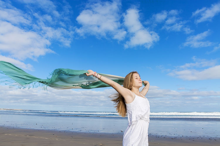 Woman holding shawl while standing at beach against sky