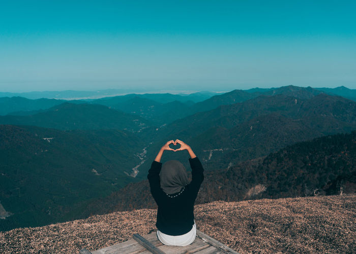 Rear view of woman making heart shape with hands while sitting on mountain 