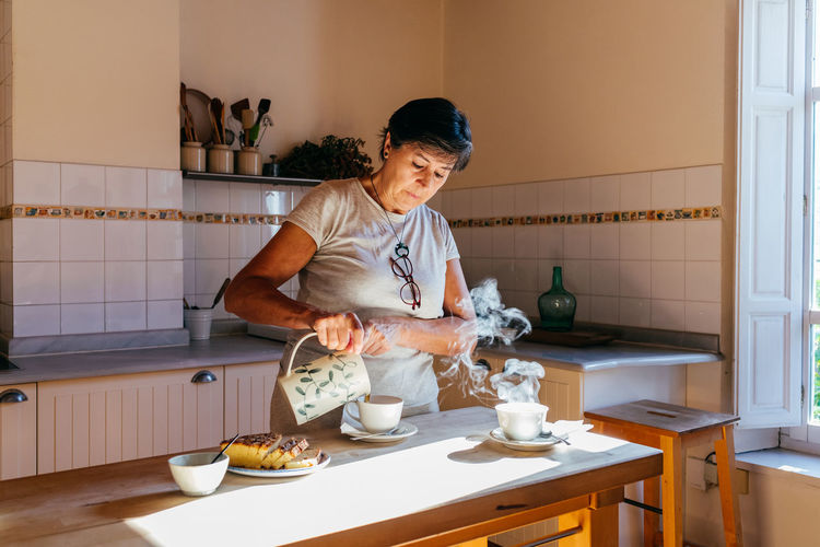 Mature female pouring hot tea from ceramic teapot into cup at table with homemade cake in cozy kitchen with wooden furniture in sunlight