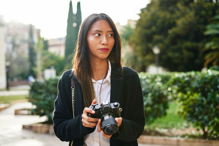Focused hispanic female in stylish outfit taking photo on modern photo camera in hands standing on street with green plants
