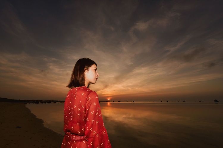 Side view of woman standing at beach against sky during sunset