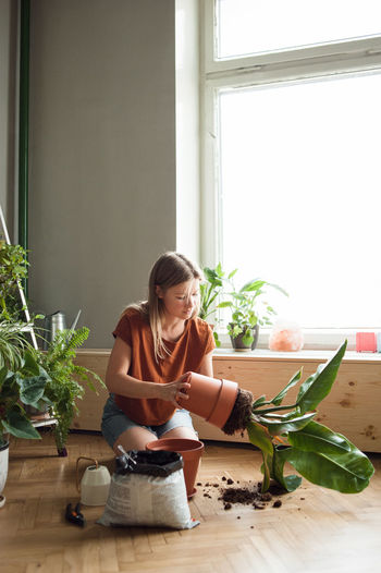 Woman takes plant of a pot, kneeling on floor with soil, garden tools.
