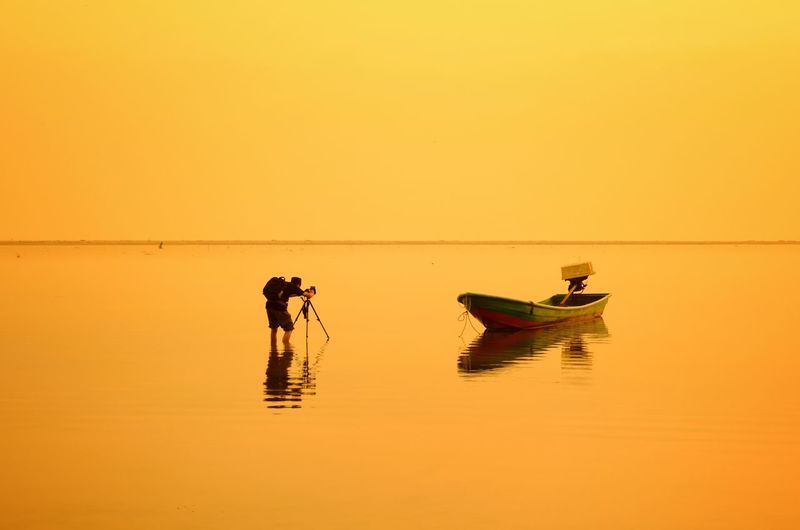 Man photographing boat moored in sea against clear sky during sunset