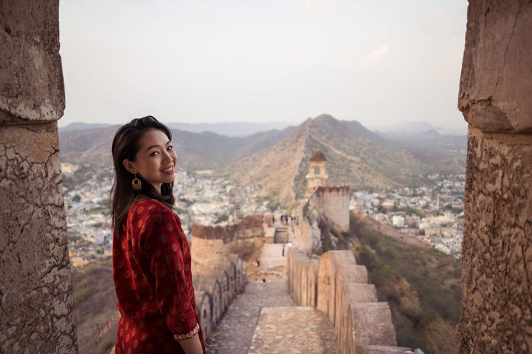 Side view of cheerful asian woman smiling and looking at camera over shoulder while visiting old amer fort viewpoint in mountains of jaipur, india
