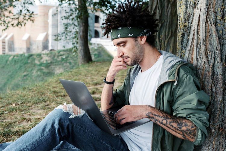 Side view of young man using laptop while sitting by tree trunk in park