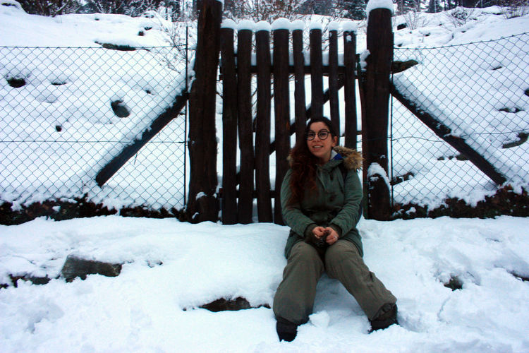 Girl in winter outfit sitting in front of a wooden fence among the white snow in daylight in tuscany