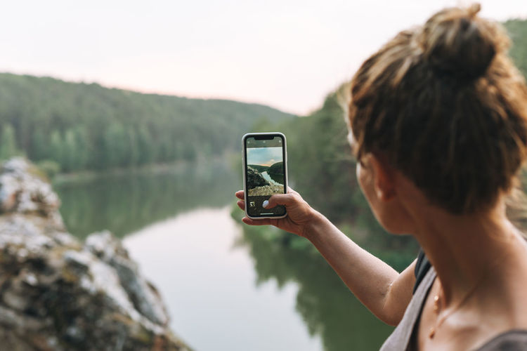 Young woman with backpack taking photo of beautiful view of mountains and calm river on mobile phone