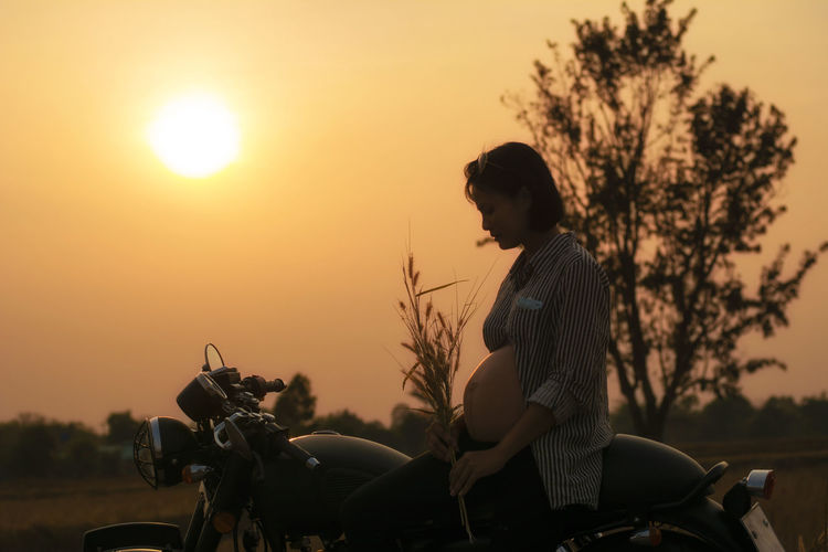 Side view of pregnant woman sitting over motorcycle against sky during sunset