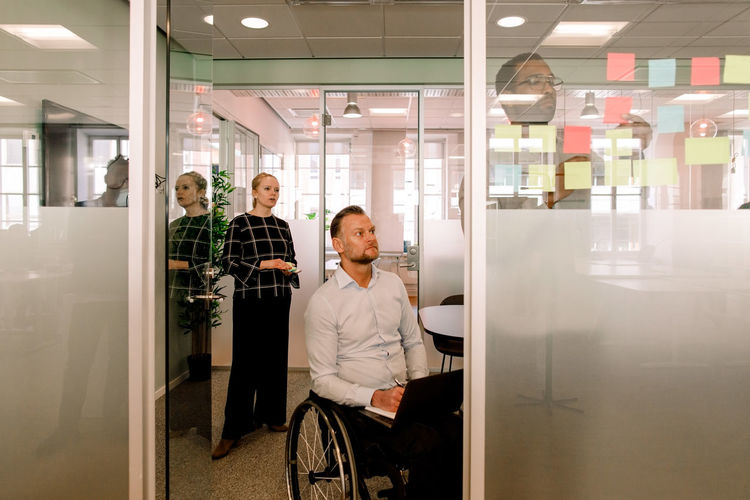 Colleagues looking at businessman sticking adhesive notes on glass wall in board room
