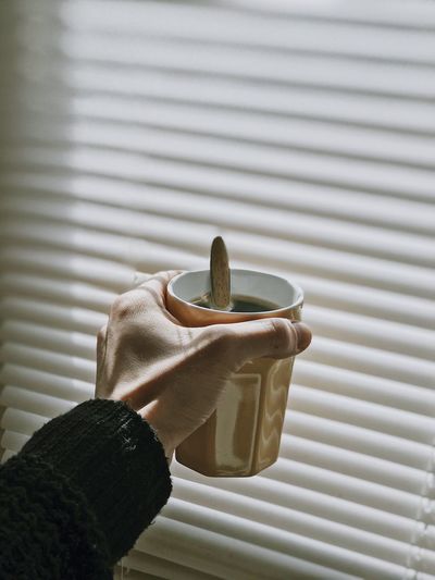 Close-up of hand holding tea in mug by blinds