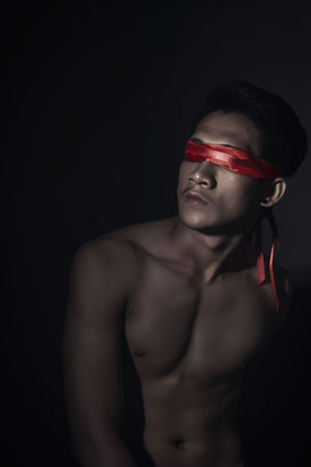 Shirtless man blindfolded with red ribbons in darkroom