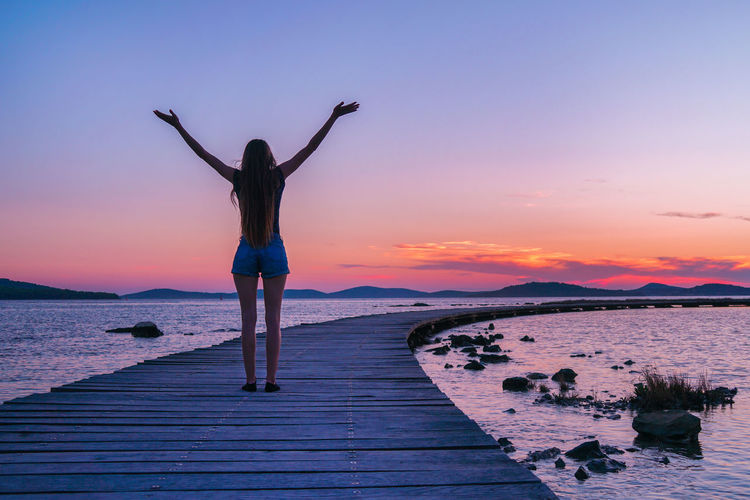 Rear view of woman standing at sea shore against sky during sunset.