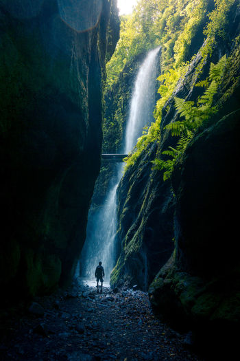 Back view of unrecognizable person walking of spectacular scenery of waterfall in long exposure in woods with green plants in highland area