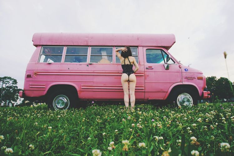 Rear view of young woman in bikini standing by car on grassy field against clear sky