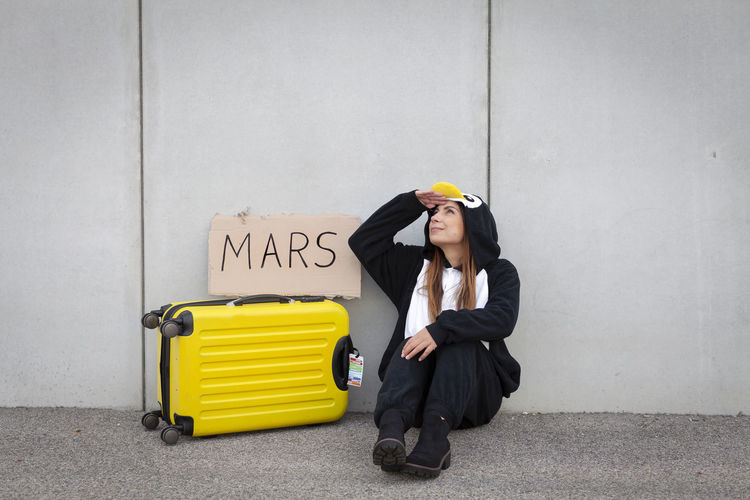 Woman sitting by luggage with text written on cardboard against wall