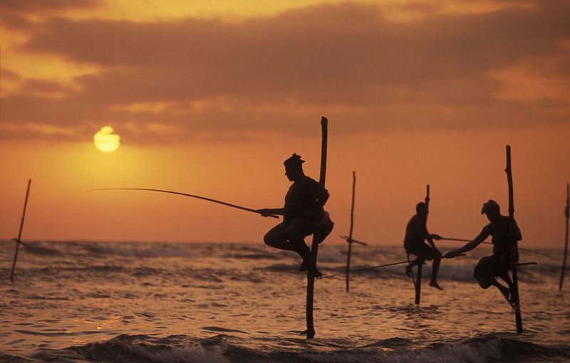 Side view of silhouette man and woman fishing in sea from wooden posts at sunset