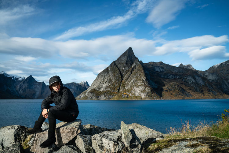 Full length of man in warm clothing sitting on rock by lake against mountain