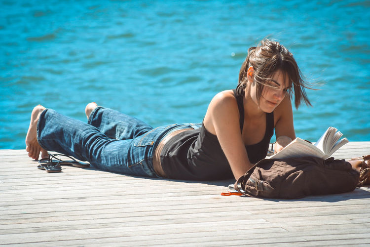 Young woman reading book while lying on jetty over sea