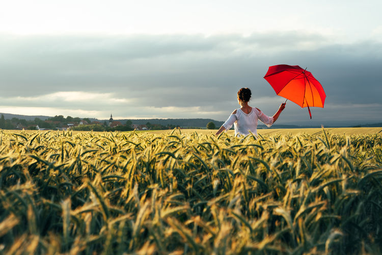 Woman standing  with a red umbrella on field against sky and enjoying the last sunbeams