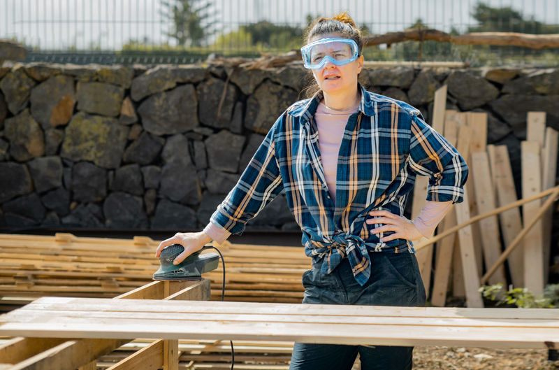 Serious female artisan in protective glasses using special electric orbital sander to smoothen wooden planks while working in countryside on summer day