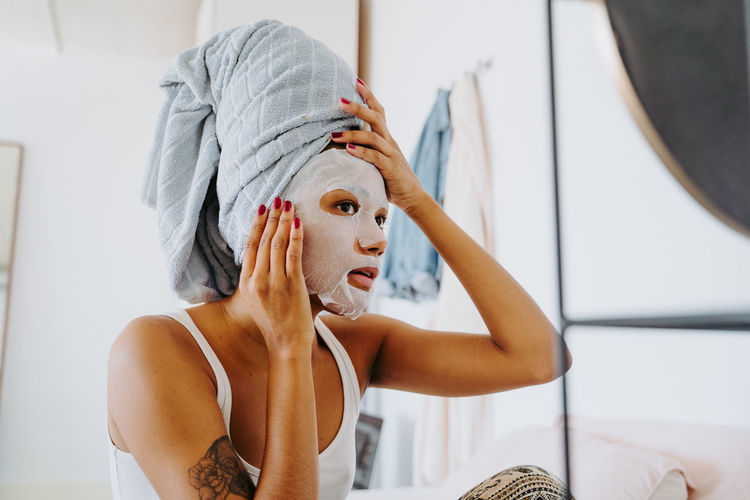 Young female in mirror applying skincare moisturizing cloth mask on face during home beauty procedure