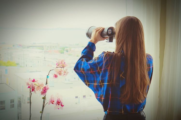 Rear view of woman looking with binoculars through glass window at home