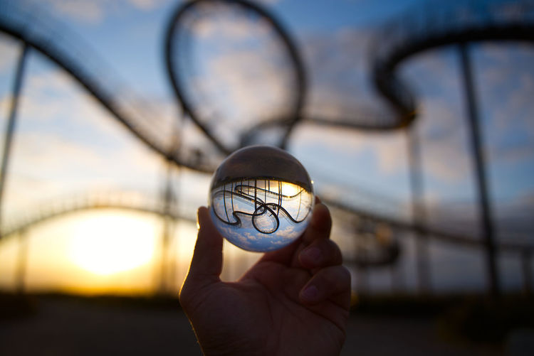 Close-up of hand holding crystal ball against roller coaster during sunset