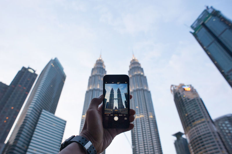 Cropped hand of man photographing petronas towers against sky
