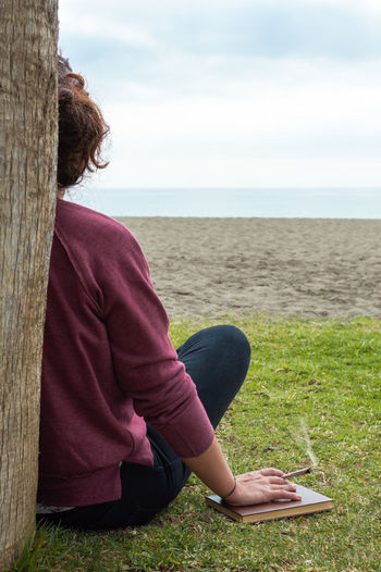 Midsection of man sitting on grass by sea against sky