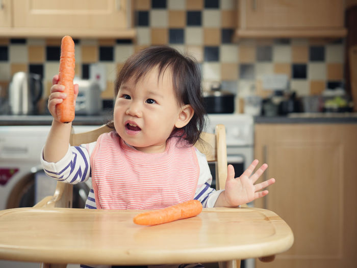 Baby girl with carrots sitting on high chair at home