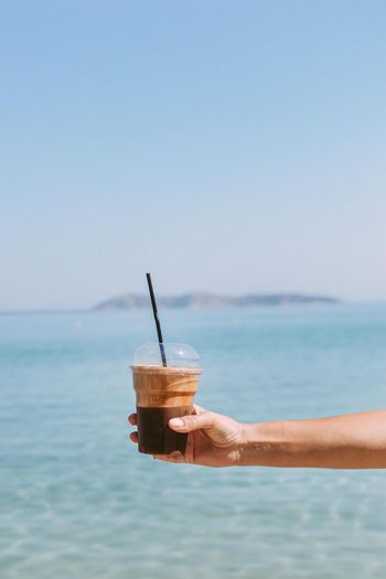 Close-up of hand holding drink by sea against sky