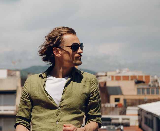 Handsome european man in sunglasses with long hair, standing outdoors and looking to the side.