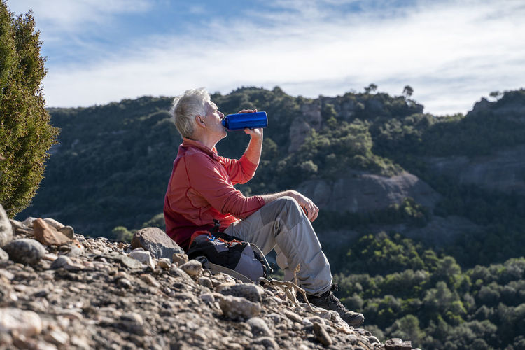 Male hiker drinking water while sitting on mountain at sant llorenc del munt i l'obac, catalonia, spain