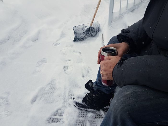 Low section of man sitting on steps, holding cup surrounded by snow and shovel