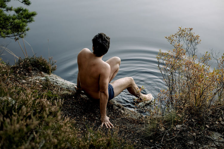 Shirtless man sitting near lake with legs dipped in water