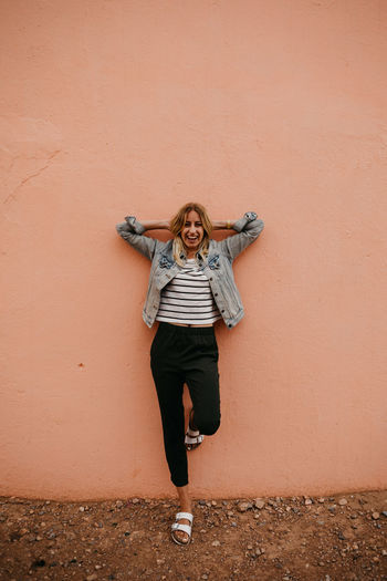 Portrait of happy young woman with hands behind head standing against wall