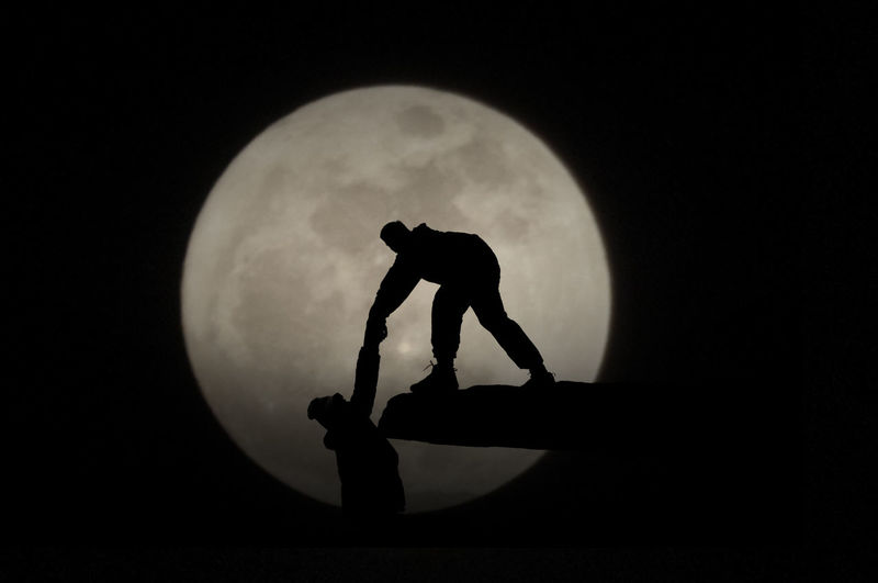 Silhouette person helping friend to climb on rock against moon at night