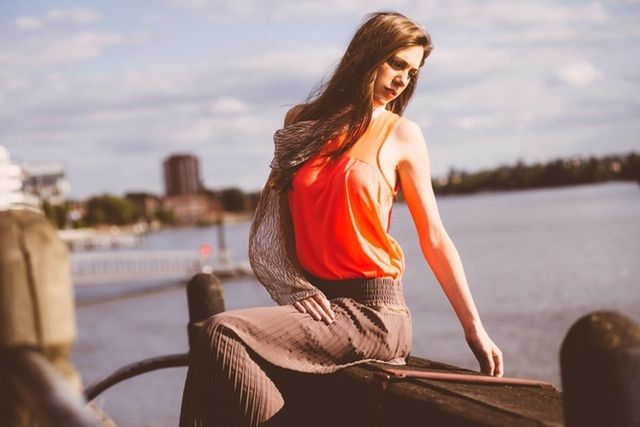 Beautiful woman sitting on retaining wall by river against sky