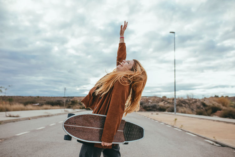 Side view of young female skater with long blond hair in trendy outfit standing on asphalt road with cruiser skateboard in hand against cloudy sky in countryside