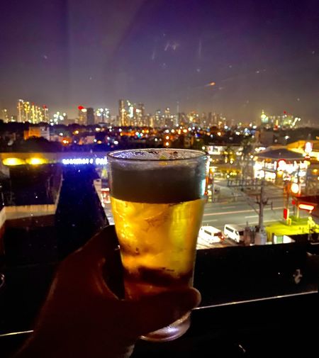Close-up of hand holding beer glass against cityscape at night