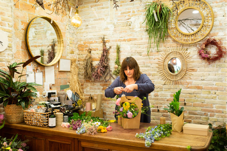 Adult female florist tying paper bag with flowers while standing behind counter and working in floral store