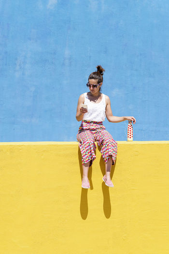 Trendy woman sitting on yellow wall while using mobile phone