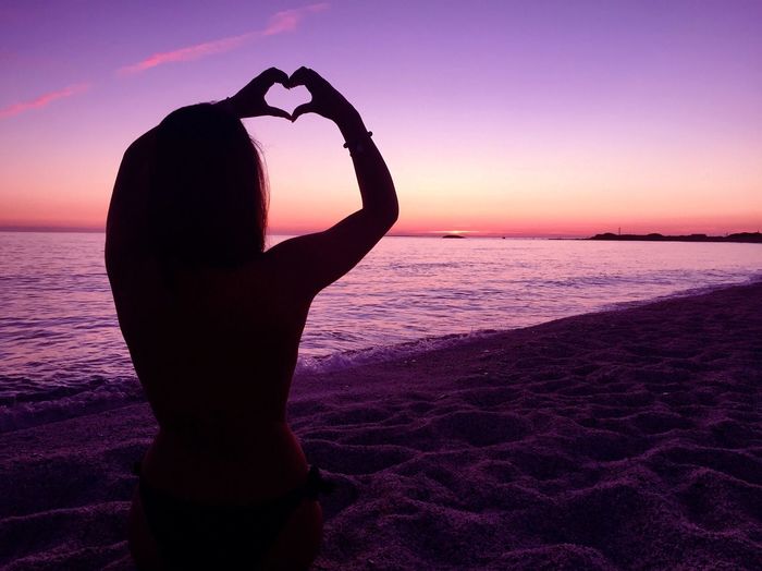 Silhouette woman making heart shape while standing at beach against sky during sunset
