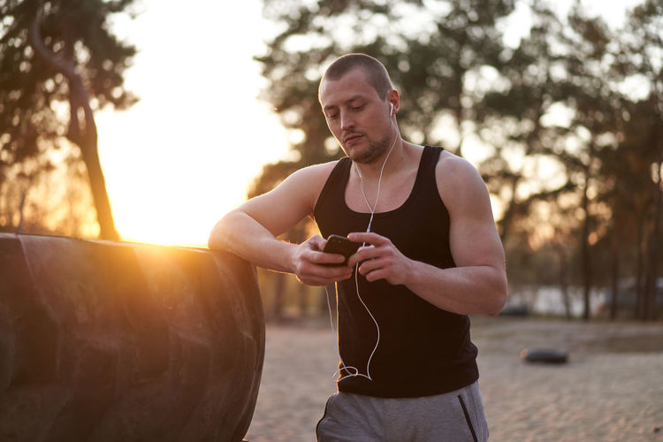 Young man using mobile phone while standing by tire at sunset