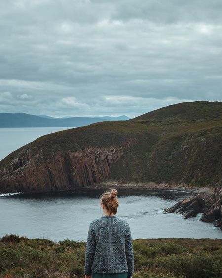 Rear view of woman looking at sea by mountain against cloudy sky