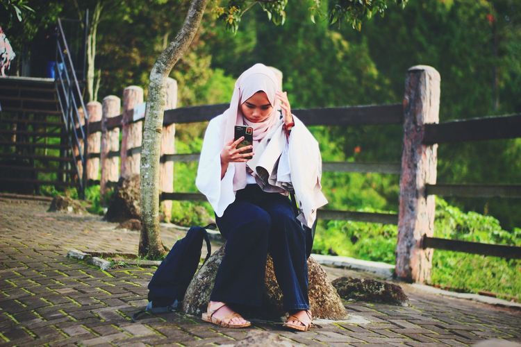 Woman wearing hijab taking selfie with smart phone while sitting in park