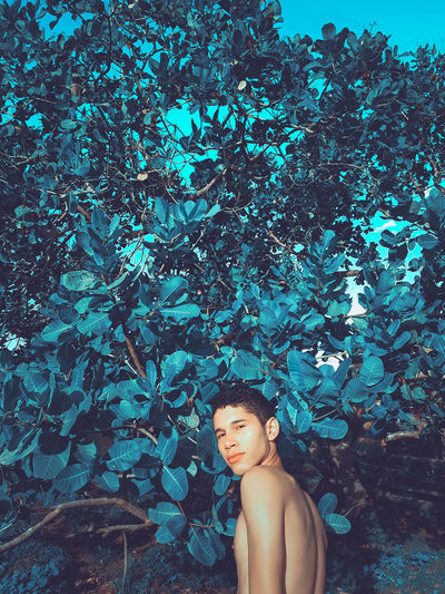 Portrait of shirtless man standing against tree